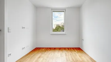 Helles, leeres Zimmer mit Parkettboden und einem Fenster, das einen Blick ins Grüne bietet.