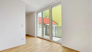 Heller, leerer Raum mit Holzboden und großen Fenstern, die auf einen Innenhof blicken.