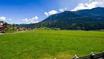 Weitläufige grüne Wiesen und Berge unter blauem Himmel, umgeben von ländlicher Bebauung.