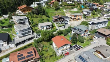 Luftaufnahme einer Wohnsiedlung am Hang mit vielen Häusern, umgeben von Wald und Grünflächen.