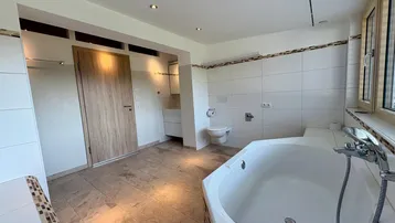 Geräumiges Badezimmer mit Eckbadewanne, WC und Fenster, hell gefliest mit Holztür und Einbaustrahlern.