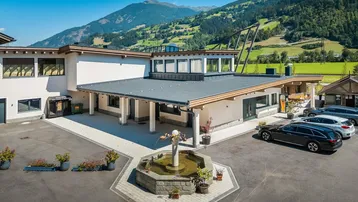 Modernes Gebäude mit großem Vorplatz, Springbrunnen und geparkten Autos vor einer malerischen Bergkulisse.