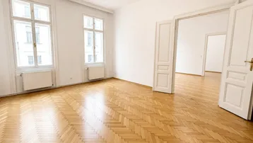 Helles Zimmer mit Fischgrätparkett, hohen Fenstern und Zugang zu weiteren Räumen.