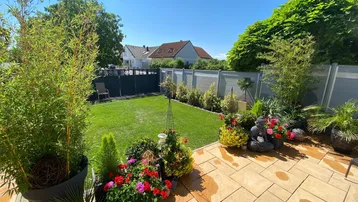 Sonnige Terrasse mit gepflastertem Bereich, üppigen Pflanzen und gepflegtem Rasen im Garten.