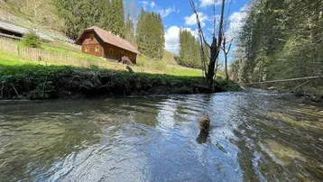 Traditionelles Holzhaus am Flussufer, umgeben von grünen Hügeln und Wäldern unter blauem Himmel.