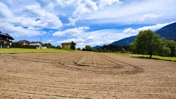 Weitläufiges Ackerland mit Häusern im Hintergrund und majestätischen Bergen unter blauem Himmel.
