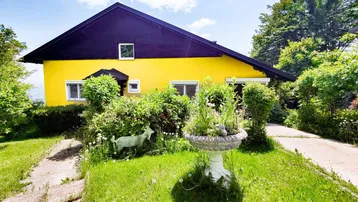 Gelbes Haus mit dunklem Dach, umgeben von viel Grün und einem kleinen Vorgarten.
