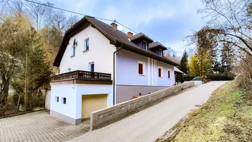 Ansicht eines Einfamilienhauses mit Balkon und Auffahrt, umgeben von grüner Natur und Bäumen.