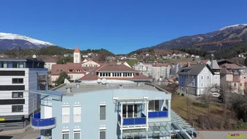 Modernes Gebäude mit blauer Fassade und Balkonen, umgeben von einer malerischen Berglandschaft und einem Dorf.