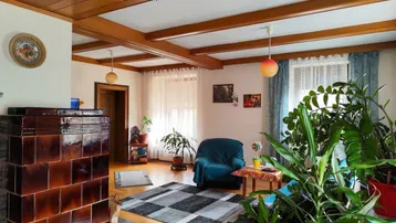 Gemütliches Wohnzimmer mit traditionellem Kachelofen, Holzbalkendecke und vielen Pflanzen.