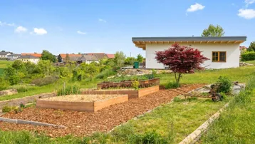 Gartenbereich mit Hochbeeten und einem kleinen Nebengebäude in ländlicher Umgebung unter blauem Himmel.