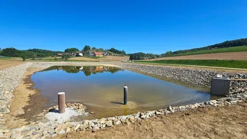 Blick auf einen Teich mit sichtbaren Entwässerungsrohren und steinernen Ufern unter klarem blauem Himmel.