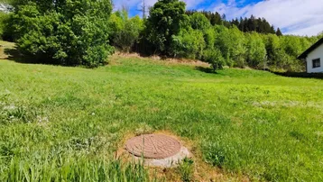 Grünes Feld mit einem Kanaldeckel im Vordergrund und bewaldeten Hügeln im Hintergrund unter blauem Himmel.