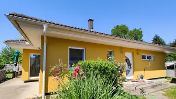 Gelbe Hausfassade mit braunem Ziegeldach und gepflegtem Vorgarten unter blauem Himmel.