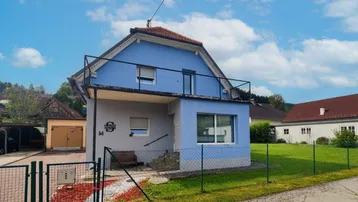 Blaues Einfamilienhaus mit Satteldach, kleinem Vorgarten und Blick auf die umliegende Landschaft.