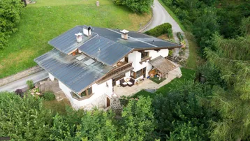 Luftaufnahme eines traditionellen Hauses mit dunklem Dach, weißer Fassade und einer Steinterrasse, umgeben von viel Grün.