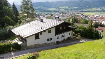Seitenansicht des Hauses mit weißer Fassade und dunklem Dach, umgeben von grünen Hügeln und einem Talblick.