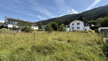 Unbebautes Grundstück mit Blick auf umliegende Häuser und die majestätische Berglandschaft unter blauem Himmel.