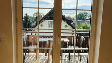 Blick durch die Fenstertüren auf einen möblierten Holzbalkon mit Sitzgelegenheiten im Freien.