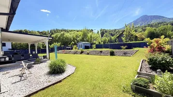 Gepflegter Garten mit großer Rasenfläche, überdachter Terrasse und Bergblick unter blauem Himmel.