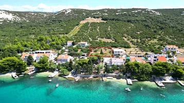 Luftaufnahme einer Küstensiedlung mit Häusern, grünen Hügeln und dem Meer, Grundstück markiert.