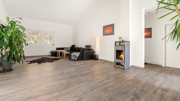 Helles Wohnzimmer mit modernem Holzofen, hohen Decken und großen Fenstern mit Blick ins Grüne.