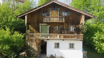 Traditionelles Holzhaus mit weißem Sockel, Holzbalkonen und grünen Fensterläden, umgeben von Bäumen.