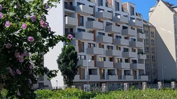 Modernes Apartmentgebäude mit vielen Balkonen, umgeben von grünen Hecken und blühenden Sträuchern.