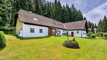 Weißes Landhaus mit braunem Ziegeldach, umgeben von einem gepflegten Rasen und dichtem Wald.
