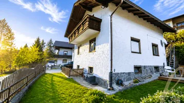 Hausfassade mit gepflegtem Garten, Rasenfläche und traditionellem Holzbalkon unter blauem Himmel.