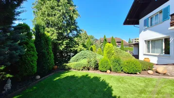 Gepflegter Gartenbereich mit Rasenfläche, Ziersträuchern und einem Teil der Hausfassade unter blauem Himmel.