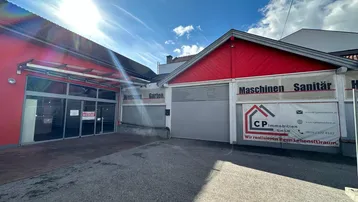 Vorderansicht eines Gewerbegebäudes mit roter Fassade und großen Schaufenstern unter blauem Himmel.