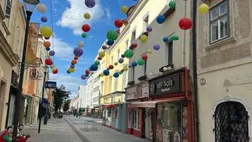 Eine belebte Einkaufsstraße mit bunten Laternen, die über die Gasse gespannt sind, unter blauem Himmel.