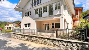 Modernes Haus mit weißer Fassade, großen Fenstern und einem gepflegten Vorgartenbereich unter blauem Himmel.