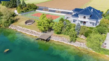 Weitläufige Ansicht des Anwesens mit Seezugang, Tennisplatz und umliegenden Feldern.