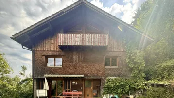Traditionelles Holzhaus mit Balkon und kleiner Terrasse im Grünen unter bewölktem Himmel.