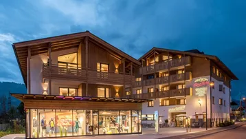 Modernes Gebäude mit Holzfassade und beleuchteten Geschäften im Erdgeschoss bei Dämmerung.