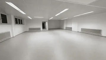 Geräumiger, leerer Büroraum mit großen Fenstern, Heizkörpern und Deckenbeleuchtung.