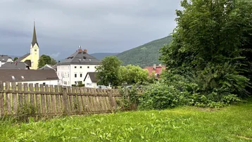 Blick über eine grüne Wiese auf ein Dorf mit Kirche und bewaldeten Bergen im Hintergrund.