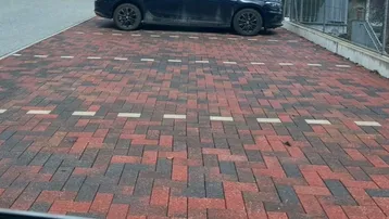 Gepflasterter Parkplatz mit Ziegelsteinen und markierten Stellplätzen, ein Auto parkt im Hintergrund auf der Straße.
