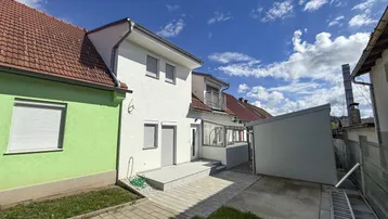 Rückseite des Hauses mit gepflastertem Bereich und angrenzendem Gebäude unter blauem Himmel.