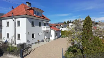Weißes Wohnhaus mit rotem Ziegeldach, Balkon und gepflasterter Auffahrt unter blauem Himmel.