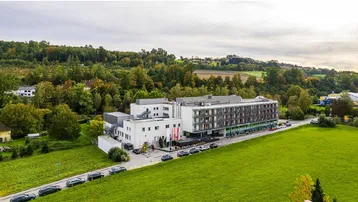 Luftaufnahme eines modernen Gebäudekomplexes, umgeben von grünen Feldern und Bäumen unter bewölktem Himmel.