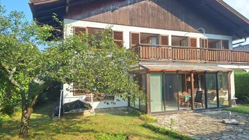 Traditionelles Haus mit Holzfassade, Wintergarten und Balkon, umgeben von einem grünen Garten.