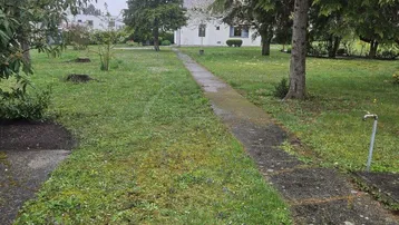 Ein langer, unbefestigter Weg führt durch den weitläufigen Garten zum weißen Haus im Hintergrund.
