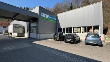 Modernes Geschäftsgebäude mit grauer Fassade und Parkplätzen vor dem Eingangsbereich unter blauem Himmel.