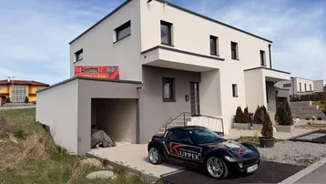 Modernes Einfamilienhaus im Bau mit Flachdach und integrierter Garage, umgeben von Grünflächen.