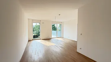 Helles, leeres Wohnzimmer mit Parkettboden und Zugang zu einem Balkon.