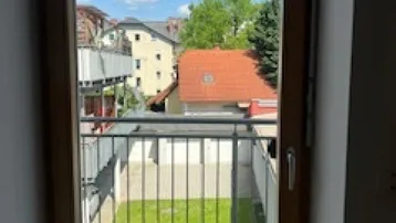 Blick vom Balkon auf einen grünen Innenhof und umliegende Gebäude unter blauem Himmel.