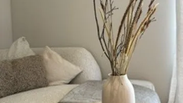 Gemütliches Wohnzimmer mit hellem Sofa, Holztischen und dekorativen Trockenblumen in einer Vase.